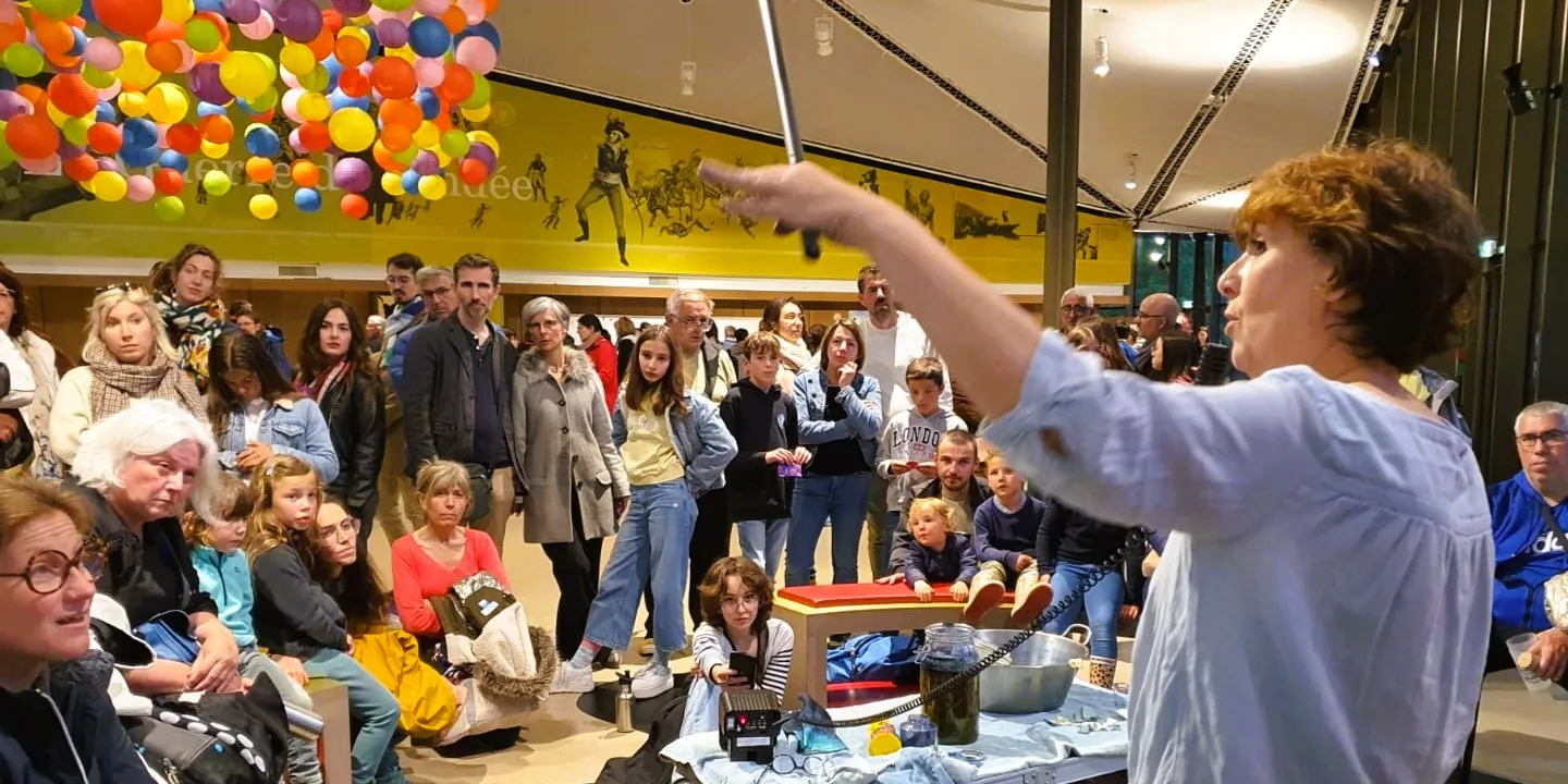 Médiation à l'historial de la Vendée Vanessa à droite en première plan de dos trois-quart, montre un objet (non visible sur l'image) avec sa main gauche tendue. Le public en face au second plan écoute les explications. EN arrière plan de le mur du musée est jaune vif et des balons multicolores sont visibles dans l'angle haut-gauche de l'image.