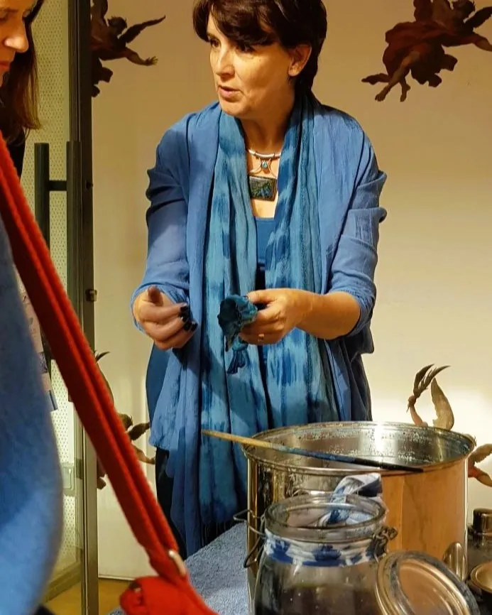 MCARMELITES 4.5 Vanessa en plan serré, habillée en bleu moyen, tient un échantillon de tissus bleu dans sa main gauche devant elle et parle avec une personne (dont on devine la présence) sur sa gauche. La lumière de fin d'après-midi est chaude.