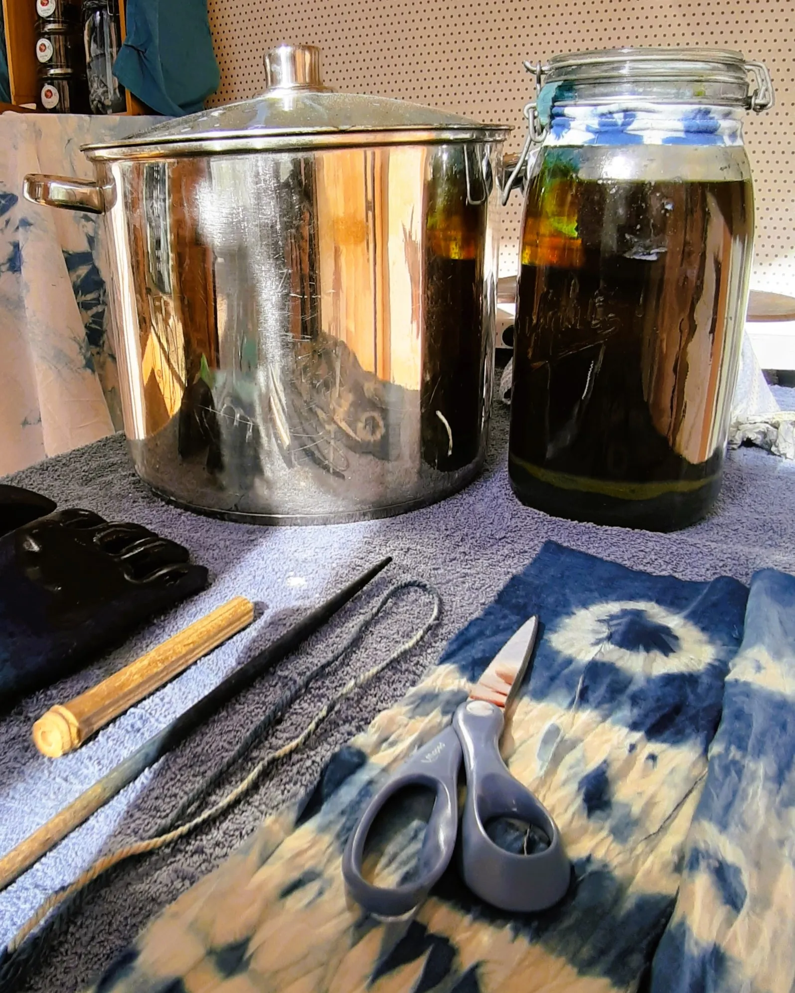 Cuves et matériel de teinture Diagonale gauche-bas et droite-haut. Au premier plan on voit des tissus shibori & indigo avec des ciseaux bleus posés dessus, on devine les gants bleus foncés à gauche. Un faitout en inox est placé à droite et une cuve d'indigo dans un bocal (couleur brun-jaune-vert) est placée à sa droite.