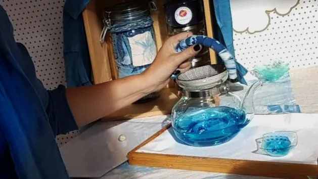 Gros plant sur le bras et la main de Vanessa qui tient l'anse de la théière en verre. Vanessa porte une chemise fluide teinte à l'indigo (moyen) ; l'anse théière est entourée de tissus blanc et bleu indigo aux motifs de shibori. Le thé visible par transparence est bleu turquoise.
